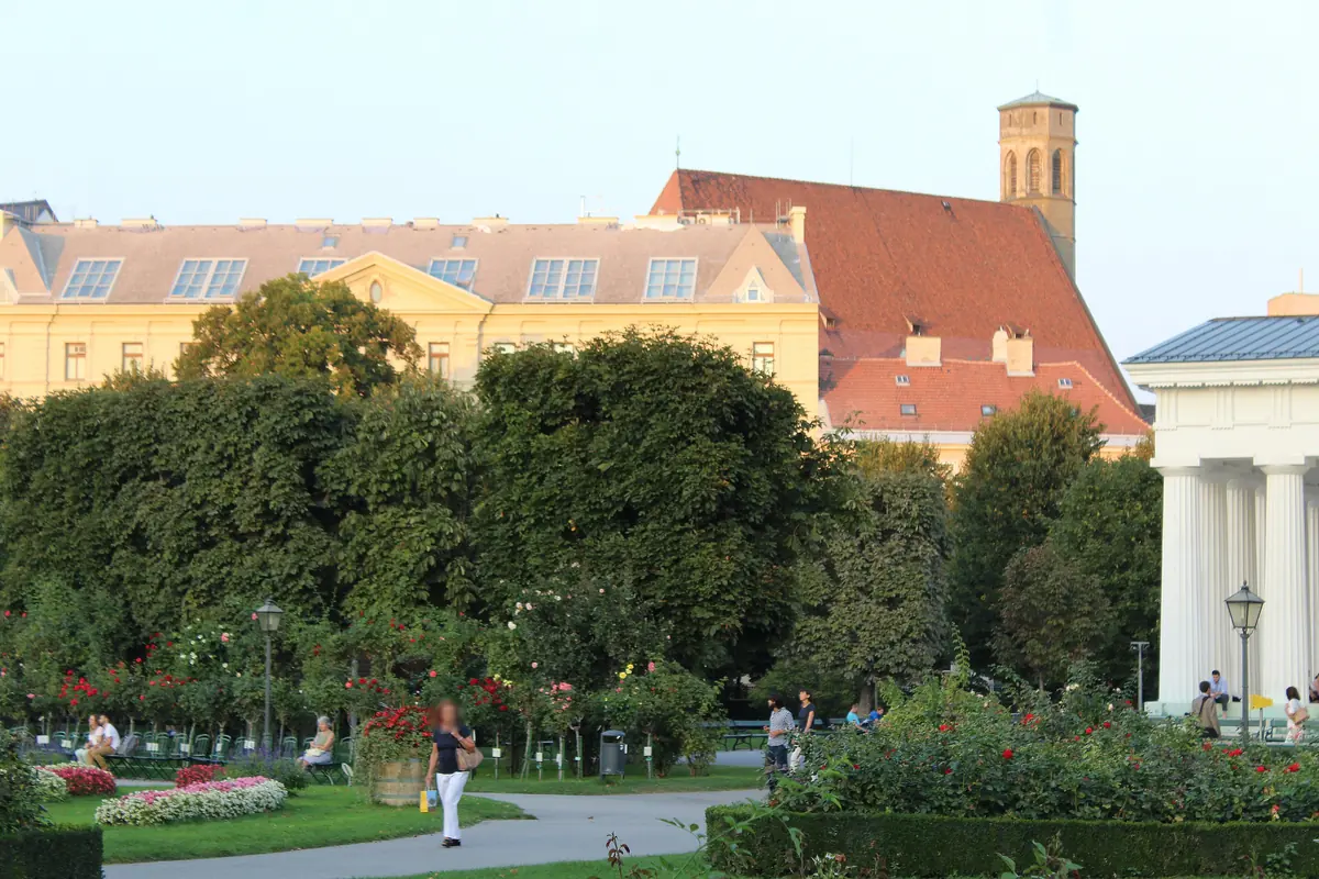 Minoritenkirche pohľadom cez zelené záhrady z Volksgartenu vo Viedni, s vežou a červenou strechou vystupujúcou za nízkou žltou budovou