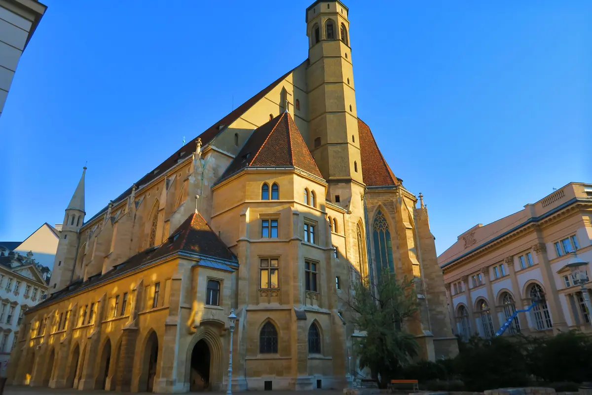 La chiesa gotica nella luce bassa della sera, con un alto tetto, una torre ottagonale e un frontone in tegole rosse accanto a un palazzo barocco.